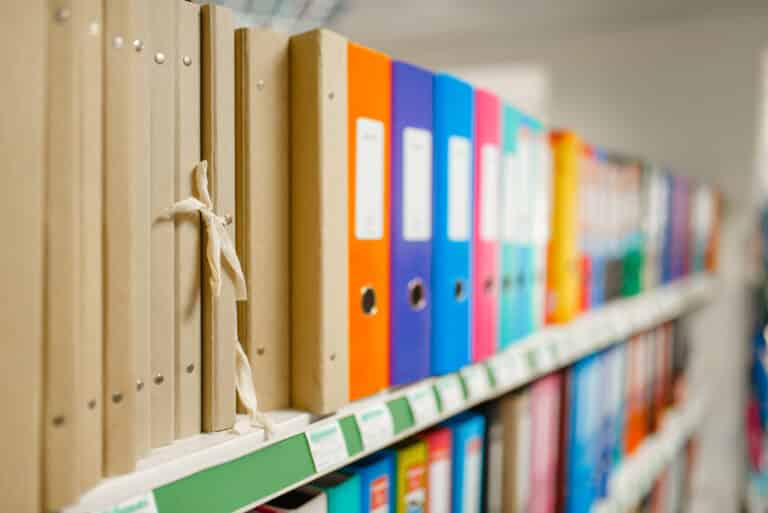 Ein Regal mit verschiedenen Ordnern in bunten Farben, darunter braune und farbige Aktenordner, die ordentlich aufgereiht sind. Im Vordergrund ist ein brauner Ordner mit einer Schnur sichtbar. Ideal für Büroorganisation und Dokumentenmanagement.