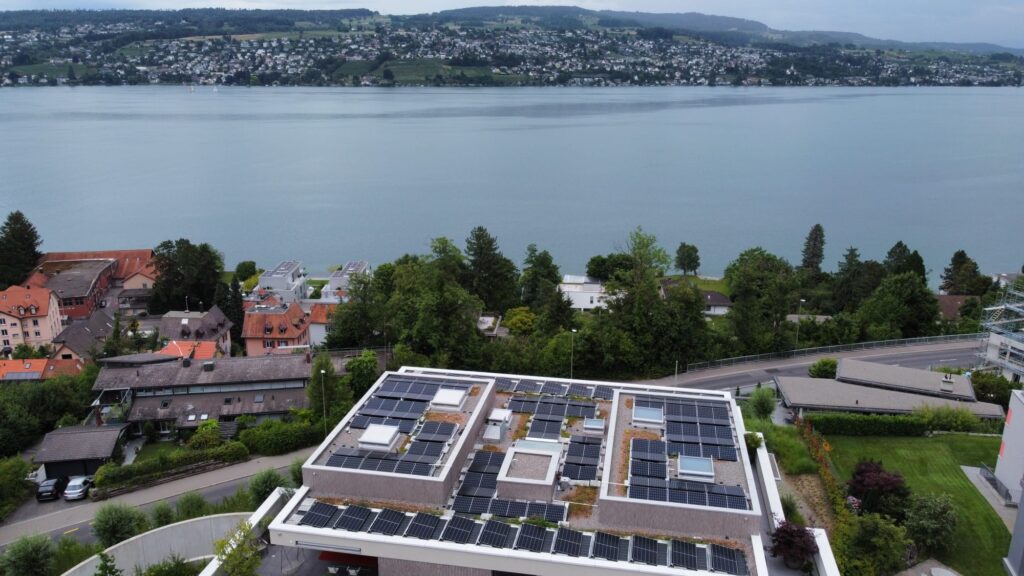Dachansicht eines modernen Gebäudes mit Solarpanelen, umgeben von grünen Bäumen und Blick auf einen See. Im Hintergrund sind Wohnhäuser und Hügel zu sehen, die eine nachhaltige Stadtentwicklung verdeutlichen.