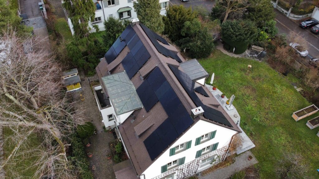Dach eines Hauses mit Solarpanelen, umgeben von einem grünen Garten und Bäumen. Die Ansicht zeigt die nachhaltige Energiequelle und die moderne Architektur des Gebäudes.
