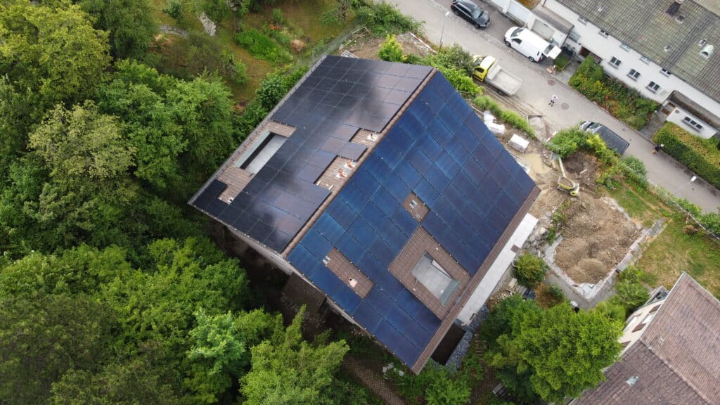 Dachansicht eines modernen Hauses mit Solarzellen in einem begrünten Wohngebiet, umgeben von Bäumen und einer Baustelle im Vordergrund.