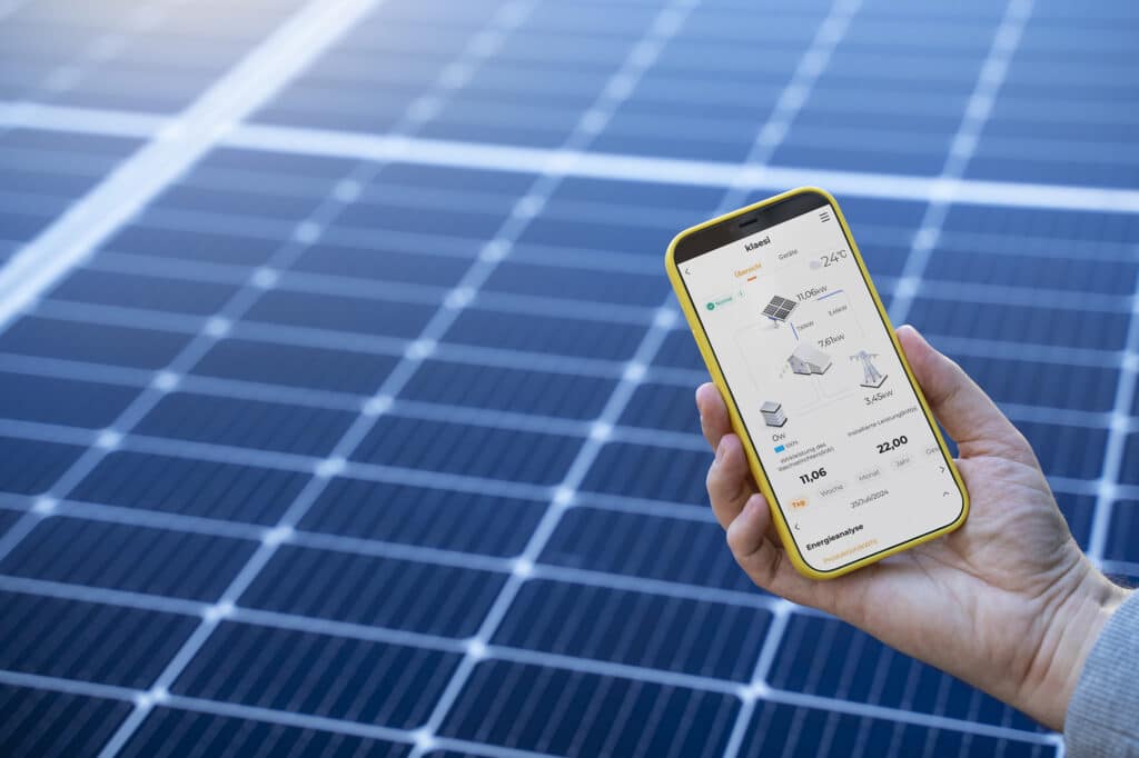 Hand, die ein Smartphone mit einer App zur Überwachung von Solaranlagen hält, vor einem Hintergrund von Solarpanels. Die App zeigt Informationen zu Energieerzeugung und -verbrauch an.