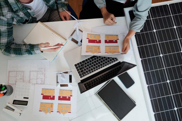 Zwei Personen arbeiten an einem Tisch mit Architekturplänen, einem Laptop und Solarpanelen. Auf dem Tisch liegen Skizzen von Gebäuden und ein Notizbuch. Die Szene zeigt eine kreative Planung im Bereich nachhaltiges Bauen.